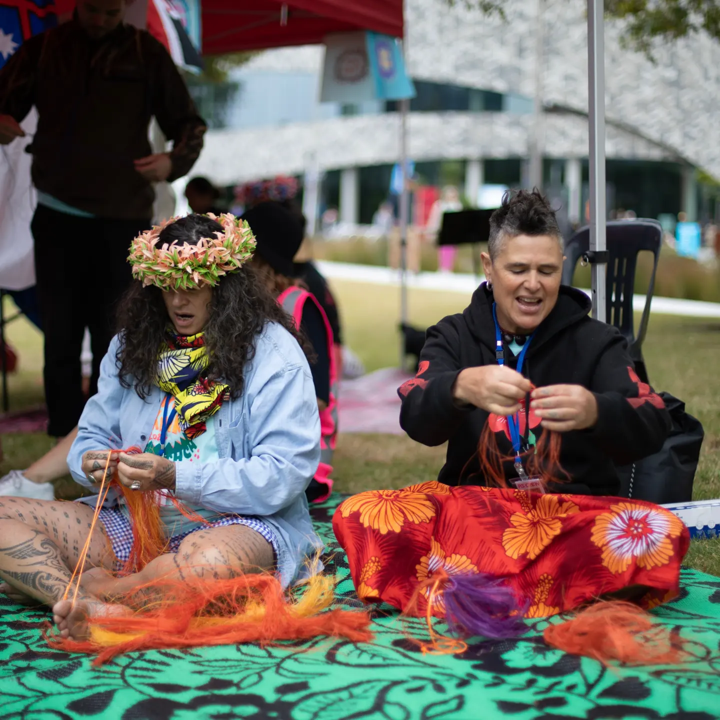 Two Pacific Sisters sitting on a green and black floral patterned mat holding orange fibers with their hands.