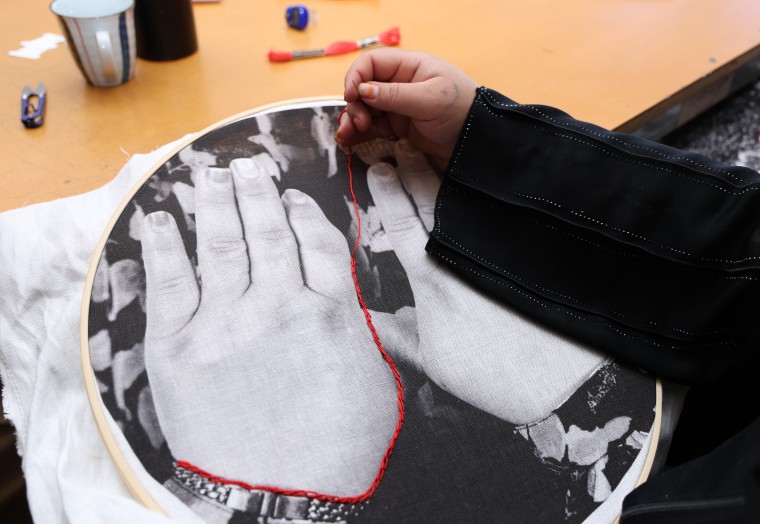 A person wearing a black top is embroidering over the top of a piece of fabric with an image printed on it. The fabric is set into a round hoop, approximately 40cm in diameter. The embroidered image is of two hands with thumbs touching and backs of hands showing. The embroidery thread is red, and the image of hands is in black and white