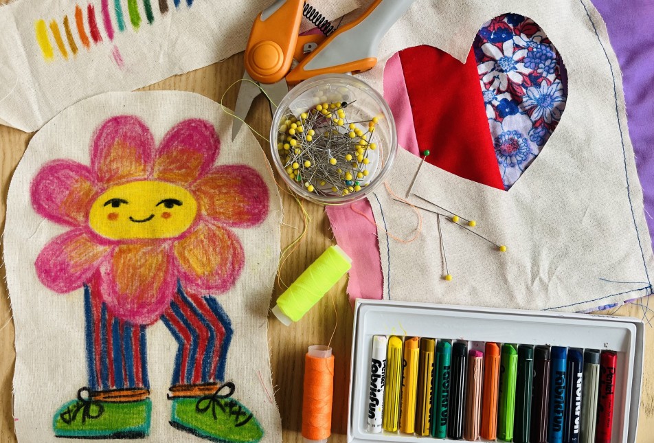 a flat lay of textile materials including coloured pastels, cotton thread in orange and yellow, a plastic container of dress makers pins, orange handled scissors and calico showing a drawing of a flower with legs and shoes