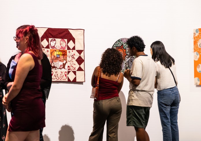 three people looking closely at an artwork on a wall. the artwork is obscured from view. next to the group of people is a white and red quilted artwork